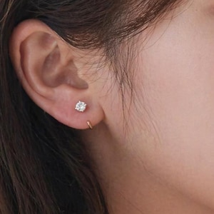 May include: A close-up of a person's ear adorned with two pieces of jewelry. One is a small, round, diamond stud earring. The other is a small, gold hoop earring. The person has brown hair and is wearing a white sweater.
