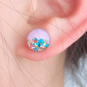 May include: A pair of purple and blue glitter stud earrings. The earrings are round and have a light purple base with a layer of colorful glitter on top.