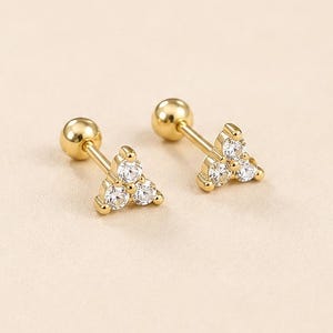 May include: A pair of gold stud earrings featuring a triangular cluster of three clear, round-cut stones. Each earring has a straight post with a spherical backing. The text LUCKY JEWUS with a small heart symbol is printed at the bottom.