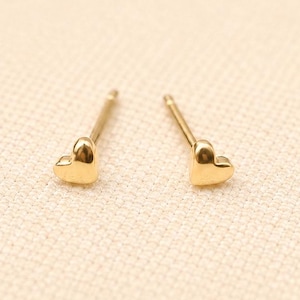 May include: A pair of small gold heart-shaped stud earrings with thin posts. The earrings feature a smooth, polished finish and a simple, classic design. The text LUCKYJEWUS is printed in small, dark letters at the bottom of the image.