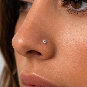 May include: Close-up of a nose with a small, sparkling diamond nose stud. The stud is set in a delicate, minimalist design. The skin tone is warm, with visible eyelashes and eyeliner.