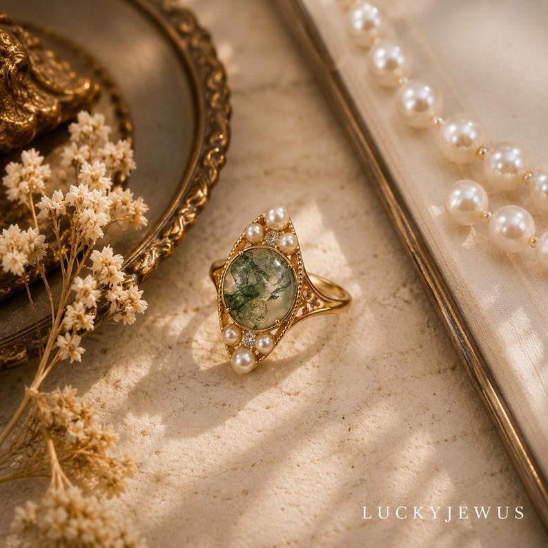 May include: A gold ring featuring a round moss agate center stone, accented by small white pearls and tiny clear stones arranged in a marquise-shaped setting. The band is thin and metallic, and the text LUCKYJEWUS is visible in the bottom right corner.