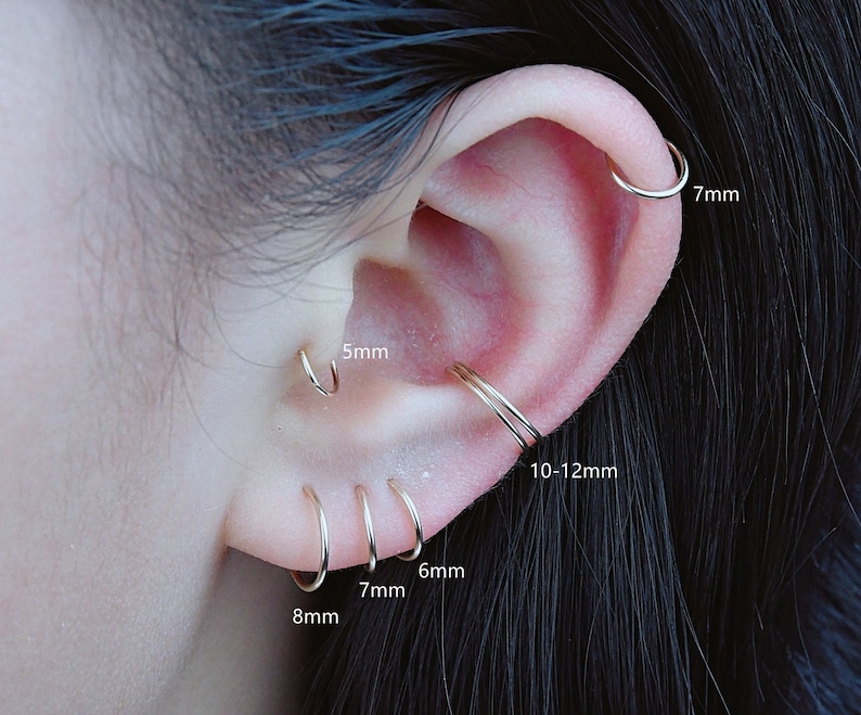May include: A close-up of a person's ear with multiple gold hoop earrings in different sizes. The earrings are labeled with their diameter in millimeters: 5mm, 6mm, 7mm, 8mm, 10-12mm, and 7mm.