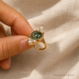 May include: A gold ring featuring a central oval moss agate cabochon surrounded by small white pearls and tiny clear stones. The metal band has a delicate beaded texture along the edges of the setting, creating a detailed and ornate appearance.