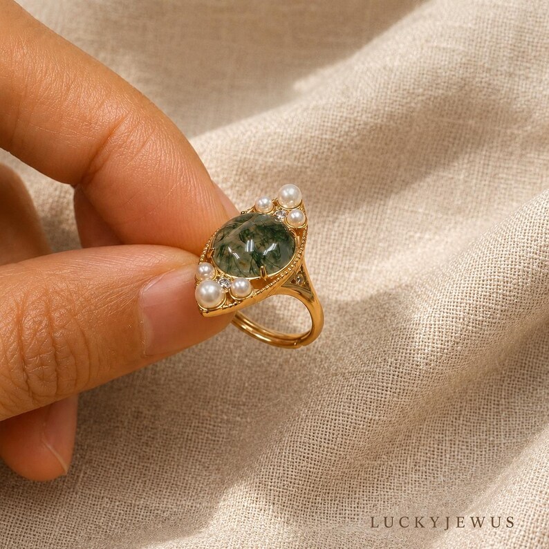 May include: A gold ring featuring a central oval moss agate cabochon surrounded by small white pearls and tiny clear stones. The metal band has a delicate beaded texture along the edges of the setting, creating a detailed and ornate appearance.