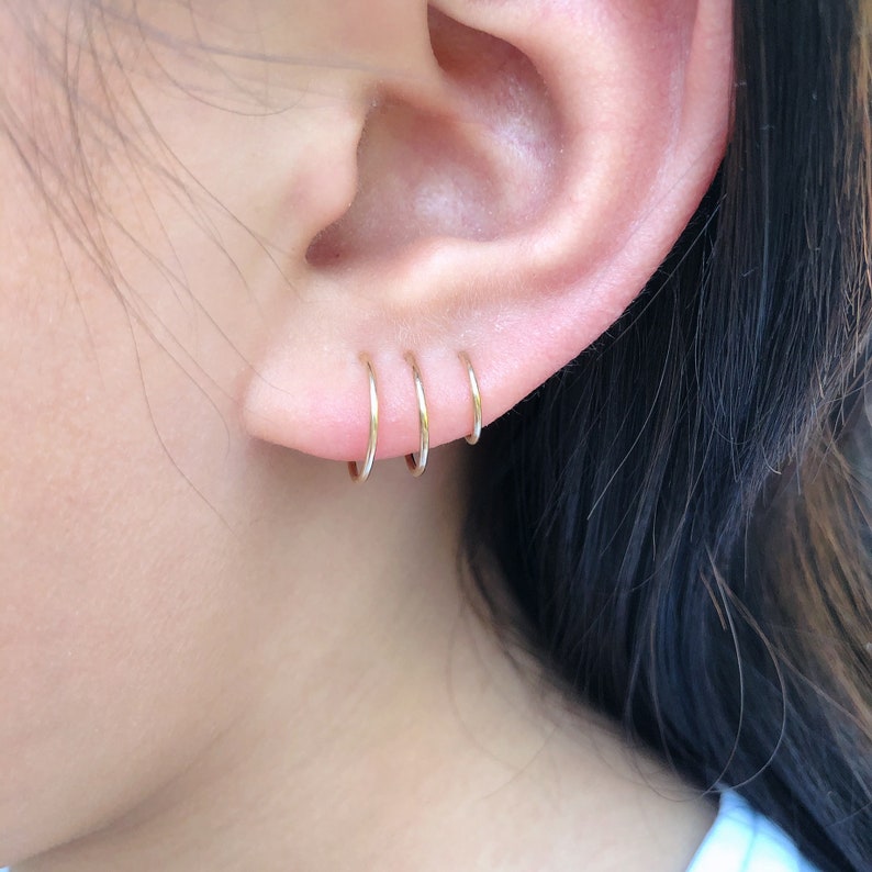 May include: Three small, gold-coloured hoop earrings are worn in a person's ear. The earrings are evenly spaced and appear to be made of a thin metal. The skin around the ear is visible, with a soft, natural tone. The background is blurred.