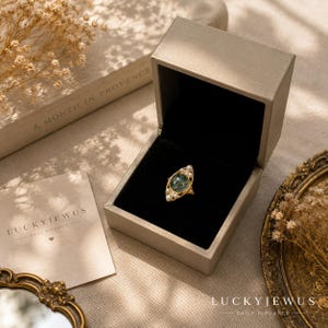 May include: A gold ring featuring a central oval moss agate stone surrounded by small white pearls, presented in an open beige jewelry box with a black interior. A card with the text LUCKYJEWUS DAILY ELEGANCE is visible next to the box.