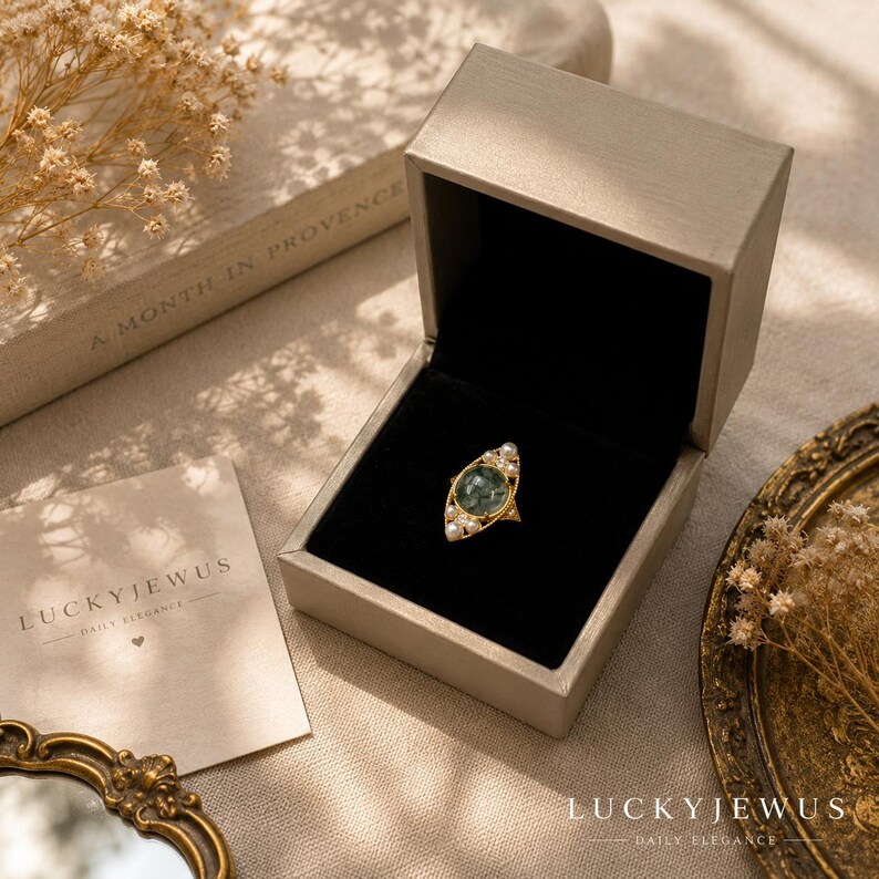 May include: A gold ring featuring a central oval moss agate stone surrounded by small white pearls, presented in an open beige jewelry box with a black interior. A card with the text LUCKYJEWUS DAILY ELEGANCE is visible next to the box.