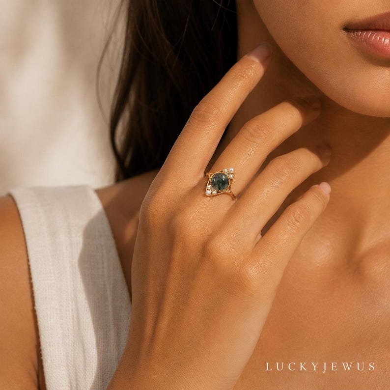 May include: A gold ring featuring a round, dark green moss agate stone set in the center, accented by small white pearls arranged along the sides of the band. The ring has a delicate, polished gold metal finish and a slender, tapered band design.