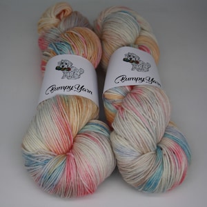 May include: Two skeins of hand-dyed yarn in a rainbow colorway with a white background. The yarn is wrapped around a white label with the text "Bumpy Yarn" and a small cartoon dog.