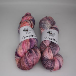 Op de afbeelding: Twee bollen handgeverfd garen in een roze, paarse en witte kleurstelling. Het garen is gewikkeld rond witte etiketten met de tekst "Bumpy Yarn" en een kleine illustratie van een hond.