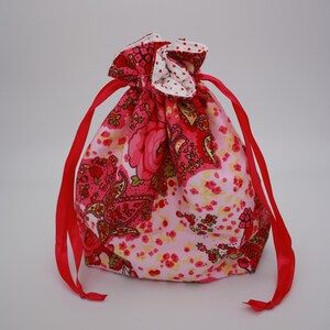 Può includere: Un sacchetto con coulisse floreale con chiusura a nastro rosso. La borsa presenta un mix di motivi floreali rosa e rossi, tra cui disegni paisley e rose. L'interno è bianco a pois rossi.