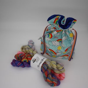 Op de afbeelding: Een set kleurrijke regenboog garenklossen met een trekkoordtas met een regenboog, wolk en zon print. De tas heeft de tekst "Love" erop. Het garen is gelabeld "Bumpy Yarn".