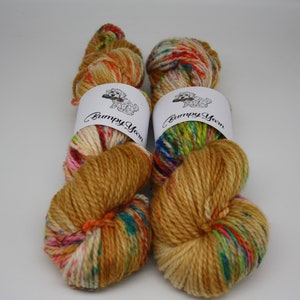 Op de afbeelding: Twee bollen garen in een gouden bruine kleur met veelkleurige vlekken. Het garen is gewikkeld rond witte etiketten met de tekst "Bumpy Yarn" en een kleine hondenillustratie.