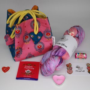 Op de afbeelding: Een roze en blauwe stoffen tas met een hartapplicatie, een streng kleurrijk garen, een theezakje, een hartvormige kaars en hartvormige versieringen. Het garen heeft een label met "Happy Yarn".