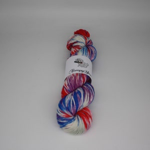 Op de afbeelding: Een bol garen in de kleuren rood, wit, blauw en paars. Het garen is gewikkeld om een wit label met de tekst "Bumpy Yarn" erop gedrukt.