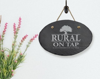 Puede incluir: Letrero ovalado de pizarra con el texto "RURAL ON TAP A NEIGHBORHOOD TAVERN" en letras blancas. El letrero está suspendido por una cuerda y cuelga de un gancho negro. Un gráfico de árbol está encima del texto. Flores artificiales en la esquina izquierda.