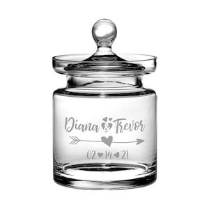 Puede incluir: Tarro de vidrio transparente con tapa, grabado con los nombres "Diana & Trevor" y la fecha "02 14 21" con corazones y una flecha.