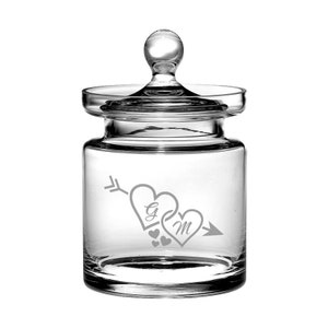 Puede incluir: Un tarro de vidrio transparente con tapa y un diseño grabado de dos corazones atravesados por una flecha. Los corazones tienen las iniciales "G" y "M" en su interior.
