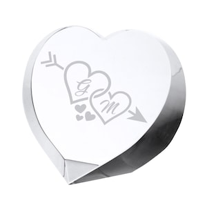 May include: A clear, heart-shaped crystal paperweight. The design features two intertwined hearts with the initials "G" and "M" and an arrow. Small hearts are also etched into the crystal. The paperweight has a faceted base.