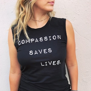 Könnte beinhalten: Schwarzes ärmelloses Top mit dem weißen Text "COMPASSION SAVES LIVES". Das Model trägt Jeansshorts und eine graue Uhr. Der Hintergrund ist eine hellfarbige Wand mit etwas Grün.