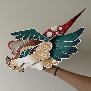 Peut inclure: Un masque de tête de dragon en papier superposé présentant une palette de couleurs sarcelle et beige avec des motifs de tourbillons blancs complexes, une corne rouge pointue et un œil vert proéminent. Le design inclut des ailes stylisées.