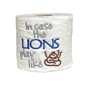 May include: A white toilet paper roll with the text "In case the LIONS play like" embroidered in gray and blue, with a brown poop emoji embroidered next to it.