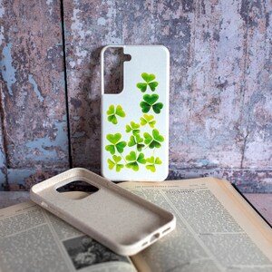 May include: A white phone case with a design of green shamrocks. The case is made of a textured material and has a cutout for the camera lens.