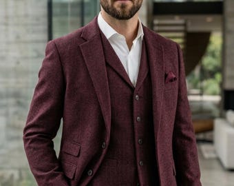 May include: A three-piece burgundy suit with a textured weave. The suit includes a blazer, vest, and trousers, paired with a white dress shirt. A pocket square and watch complete the ensemble. The suit is a classic formal wear.