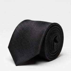 May include: A rolled black tie with a repeating geometric design. The tie is made of a dark, smooth material and is presented against a plain white backdrop. The texture and pattern of the tie are clearly visible.