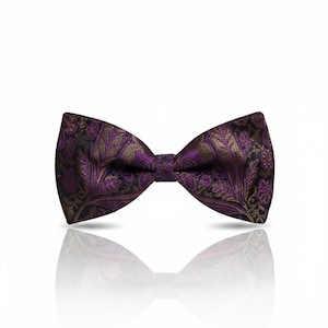 May include: A purple bow tie with a gold and black floral design. The bow tie is set against a white backdrop, with its reflection beneath. The fabric has a textured finish.