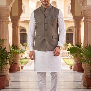 May include: A man in a traditional Indian outfit. He wears a white kurta and trousers, a brown waistcoat, and navy blue shoes. The background shows an ornate building with archways and columns.
