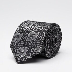 May include: Rolled black tie with a silver floral pattern. The tie is made of a smooth fabric and features a repeating design of stylized flowers and swirls. The tie is rolled up, showing the intricate pattern.