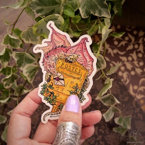 Può includere: Un adesivo raffigurante un drago appollaiato su una cassetta postale gialla con la scritta "POSTES". Il drago è rosa e arancione e la cassetta postale ha una fessura per le lettere con una lettera all'interno. Viti e foglie verdi circondano la cassetta postale.