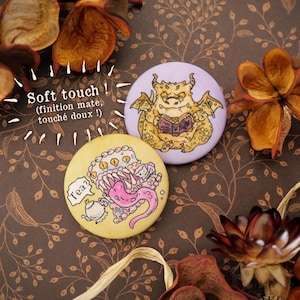 Peut inclure: Deux badges ronds et colorés avec des illustrations de monstres de dessins animés. L'un est jaune avec un monstre et une théière avec l'inscription "Tea ?". L'autre est violet avec un dragon lisant un livre. Le texte "Soft touch !" est également visible.