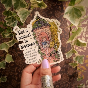 May include: A sticker with the text "Not a damsel in distress." The illustration depicts a princess reading in a tower, with a dragon. The sticker is white with a decorative border and a floral design.