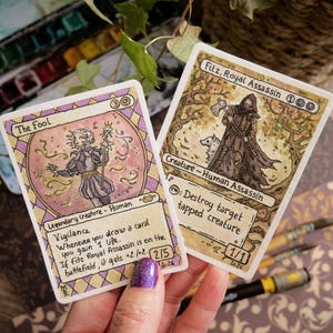 Può includere: Due carte collezionabili dipinte a mano con illustrazioni fantasy. La carta a sinistra intitolata The Fool raffigura una figura con un bastone, mentre la carta a destra Fitz, Royal Assassin mostra un personaggio incappucciato con un'ascia.