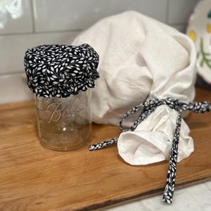 May include: A clear glass jar with a black and white leaf-patterned lid sits on a wooden surface. Beside it is a white cloth bag tied with a black and white patterned ribbon. The jar is labeled "Ball".