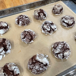 Puede incluir: Una bandeja para hornear contiene una docena de galletas de chocolate crujientes. Las galletas son de color marr&oacute;n oscuro con una capa agrietada de az&uacute;car glas. Se encuentran sobre papel de hornear dentro de una bandeja met&aacute;lica.