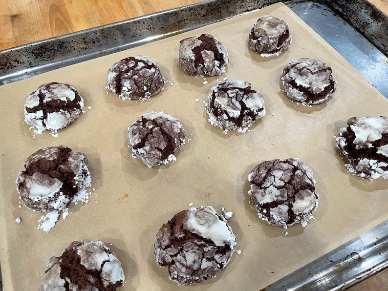 Puede incluir: Una bandeja para hornear contiene una docena de galletas de chocolate crujientes. Las galletas son de color marr&oacute;n oscuro con una capa agrietada de az&uacute;car glas. Se encuentran sobre papel de hornear dentro de una bandeja met&aacute;lica.