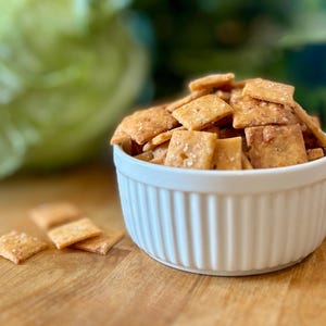 Puede incluir: Un cuenco blanco acanalado lleno de galletas cuadradas de color marr&oacute;n dorado, algunas espolvoreadas con sal. Varias galletas est&aacute;n esparcidas sobre la superficie de madera. El fondo presenta una verdura verde borrosa y vegetaci&oacute;n.