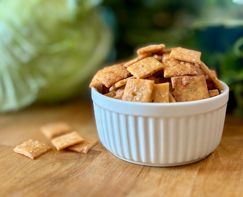 Puede incluir: Un cuenco blanco acanalado lleno de galletas cuadradas de color marr&oacute;n dorado, algunas espolvoreadas con sal. Varias galletas est&aacute;n esparcidas sobre la superficie de madera. El fondo presenta una verdura verde borrosa y vegetaci&oacute;n.