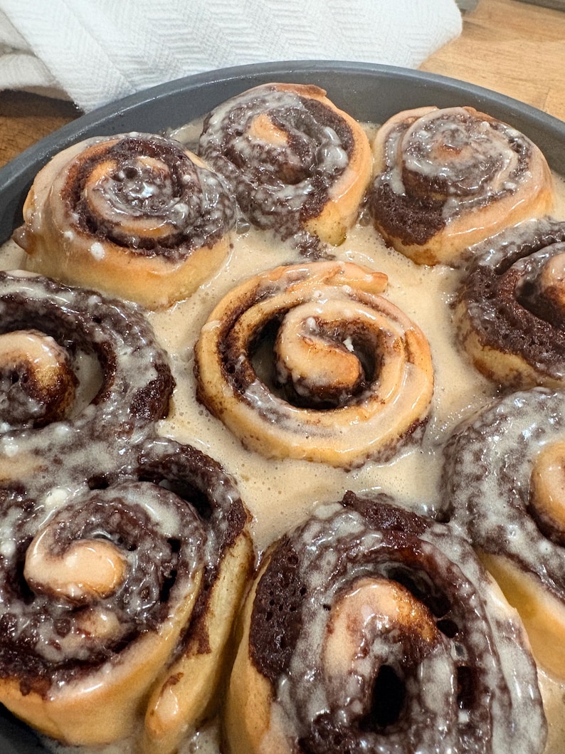 Puede incluir: Un molde circular de metal oscuro lleno de varios rollos de canela dorados. Cada rollo presenta una espiral de relleno de canela de color marr&oacute;n oscuro y est&aacute; cubierto con una capa de glaseado de az&uacute;car blanco transl&uacute;cido.