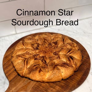 Puede incluir: Pan de masa madre con forma de estrella de canela, de color dorado, presentado sobre una tabla de madera. El pan tiene un dise&ntilde;o en forma de estrella y est&aacute; espolvoreado con canela. El texto "Cinnamon Star Sourdough Bread" es visible.