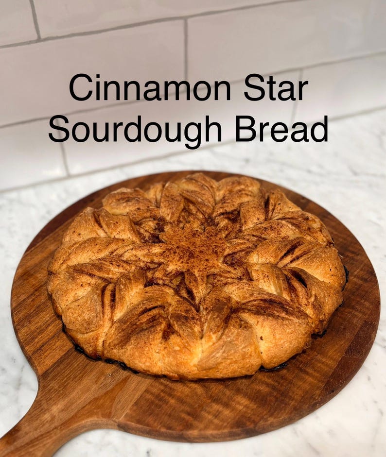 Puede incluir: Pan de masa madre con forma de estrella de canela, de color dorado, presentado sobre una tabla de madera. El pan tiene un dise&ntilde;o en forma de estrella y est&aacute; espolvoreado con canela. El texto "Cinnamon Star Sourdough Bread" es visible.