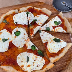 Puede incluir: Una pizza en rodajas con queso mozzarella, salsa de tomate y hojas de albahaca sobre una tabla de cortar de madera. Un cortador de pizza est&aacute; en la esquina superior derecha. La pizza est&aacute; cortada en ocho rebanadas.