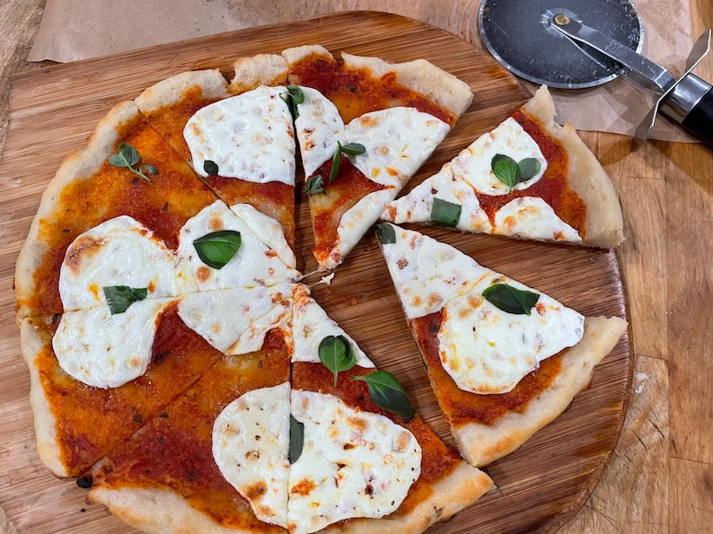 Puede incluir: Una pizza en rodajas con queso mozzarella, salsa de tomate y hojas de albahaca sobre una tabla de cortar de madera. Un cortador de pizza est&aacute; en la esquina superior derecha. La pizza est&aacute; cortada en ocho rebanadas.