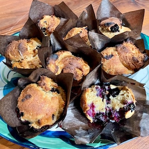 Puede incluir: Un plato de muffins de ar&aacute;ndanos reci&eacute;n horneados, cada uno en un envoltorio de papel marr&oacute;n. Los muffins tienen la parte superior dorada y est&aacute;n rellenos de jugosos ar&aacute;ndanos. El plato es azul y verde, sobre una mesa de madera.