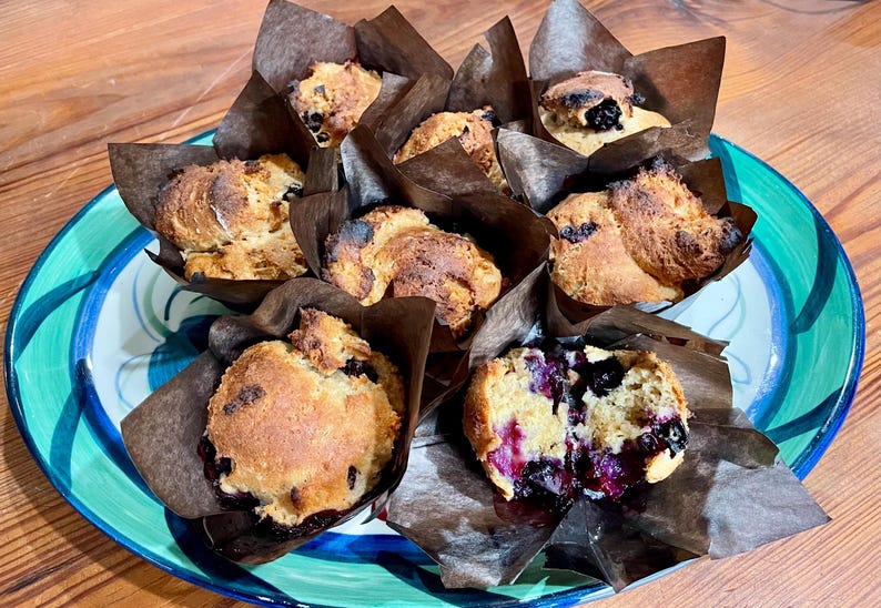 Puede incluir: Un plato de muffins de ar&aacute;ndanos reci&eacute;n horneados, cada uno en un envoltorio de papel marr&oacute;n. Los muffins tienen la parte superior dorada y est&aacute;n rellenos de jugosos ar&aacute;ndanos. El plato es azul y verde, sobre una mesa de madera.