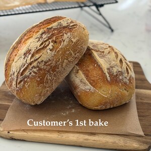 Puede incluir: Dos hogazas de pan de masa madre ovaladas, de corteza dorada y parte superior marcada, sobre un trozo de papel de horno marr&oacute;n en una tabla de cortar de madera. El texto "Customer's 1st bake" est&aacute; impreso en blanco sobre el papel.